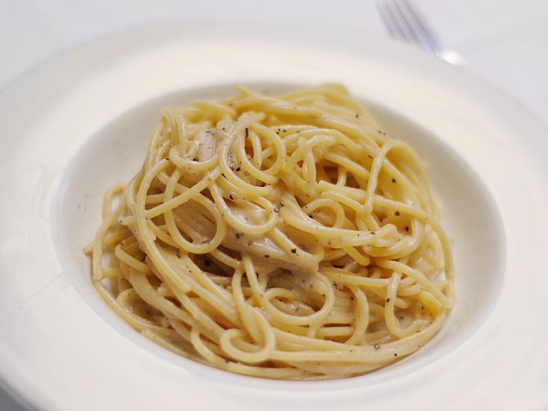 Ninho de massa longa em prato branco, coberto por creme de queijo pecorino e pimenta.