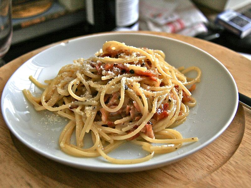 Spaghetti alla carbonara enrolados num prato raso, com pedaços de guanciale e pimenta moída por cima.