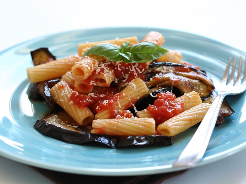 Rigatoni com molho de tomate e fatias de berinjela frita, servidos em prato azul-claro.