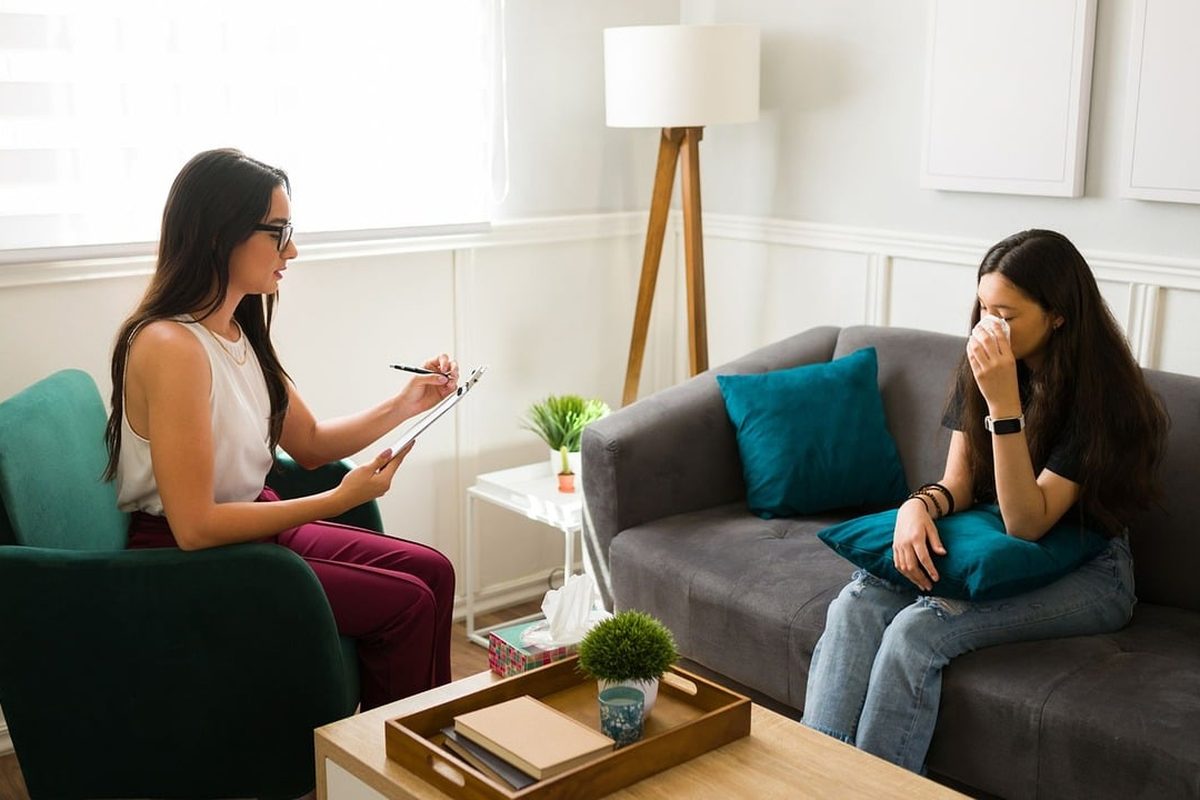 Psicóloga e paciente conversando em um sofá, com caderno de anotações e plantas na sala.
